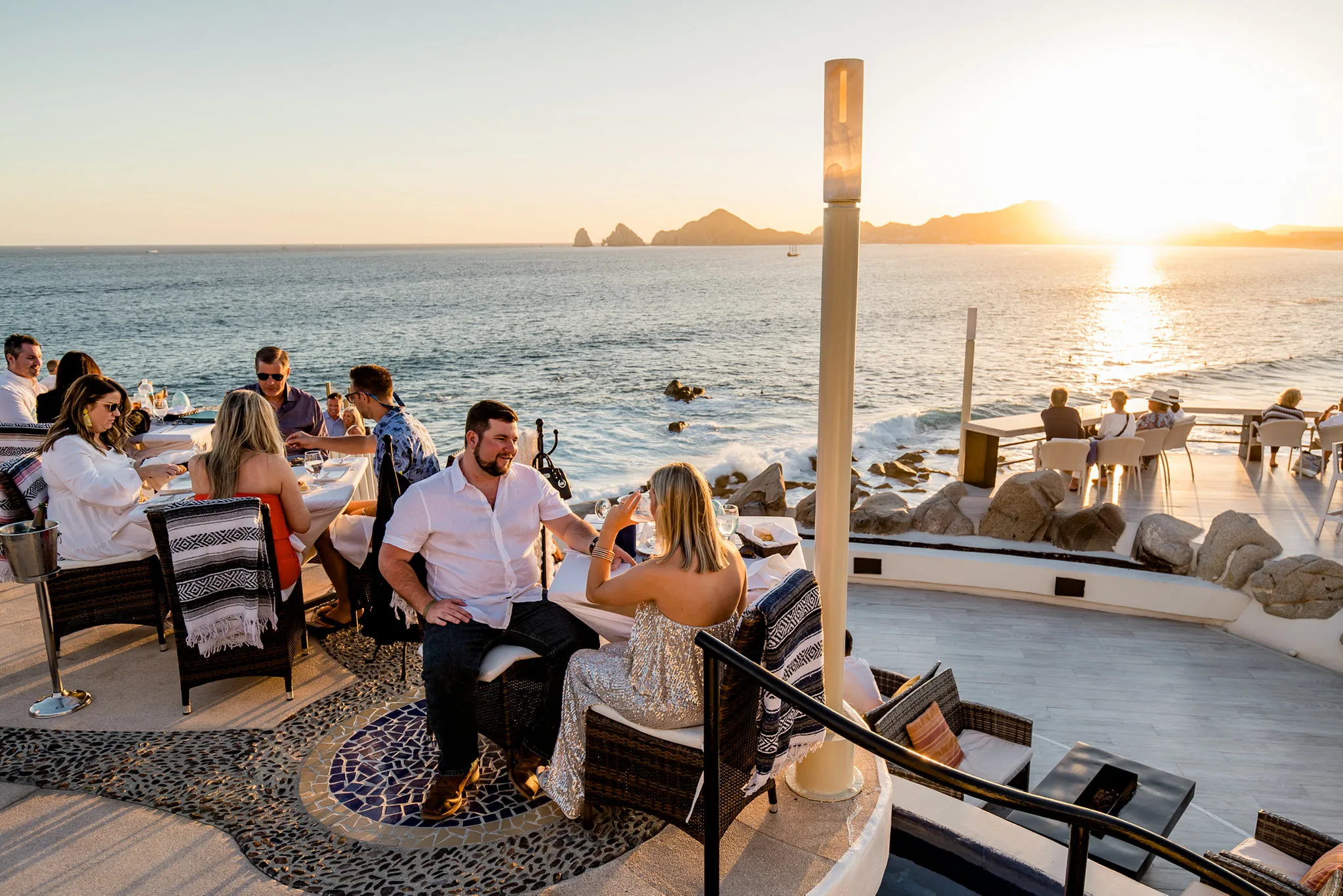 Invitados cenando en una terraza sobre el mar al atardecer, con Land’s End en el horizonte